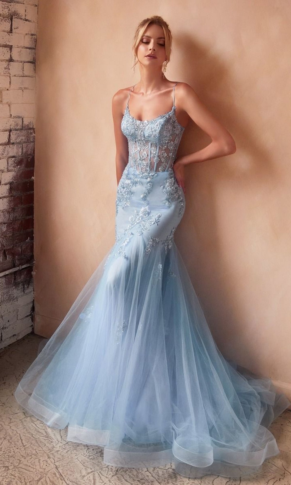 A woman wears the Long Pastel Floral Mermaid Prom Dress D145—a light blue, sleeveless gown with embroidered florals and a sheer bodice—standing against a textured wall.